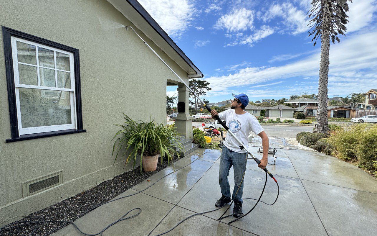 Man power washing house exterior under blue sky.